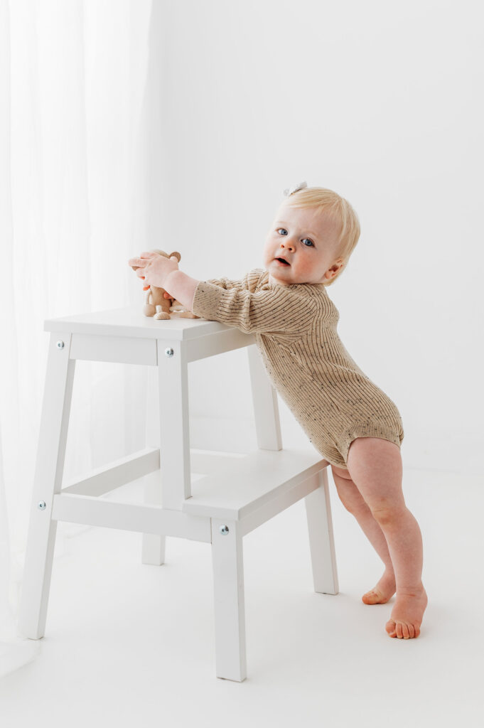 Baby girl in beige romper, standing against a white stool
