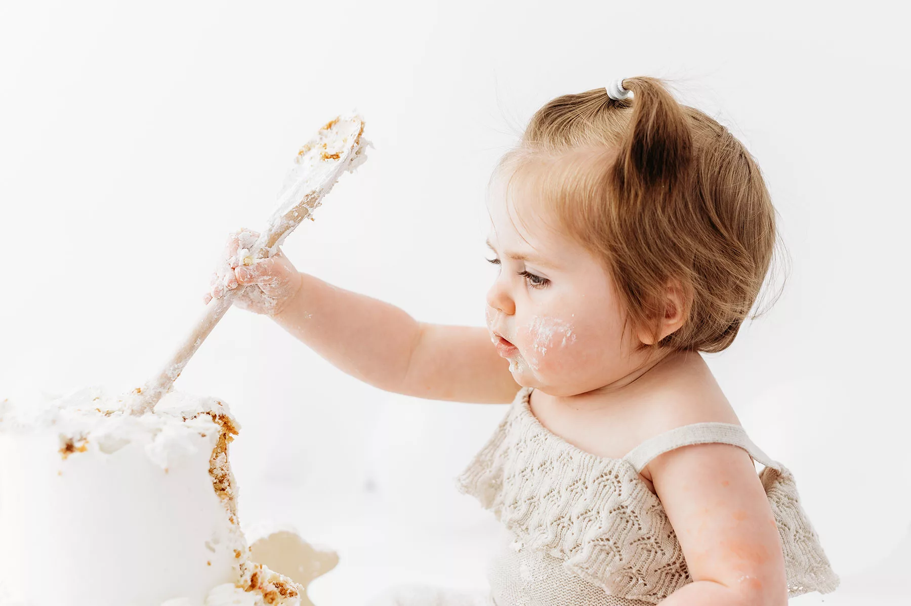 Kent cake smash photoshoot- baby girl in beige romper holding wooden spoon.
